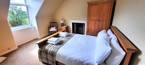 Ein Schlafzimmer mit Bett, Nachttisch, Stuhl, Kleiderschrank, Kamin und Fenster mit Blick auf Bäume.