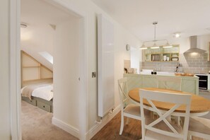 Ein Schlafzimmer mit schrägen Wänden, ein Essbereich mit rundem Tisch und Stühlen sowie eine Küche mit integrierter Kochinsel und Pendelleuchten.