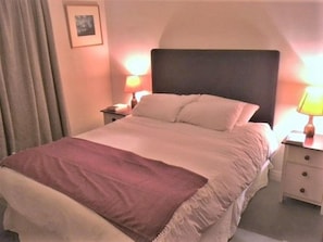 Una cama con cabecera, dos mesitas de noche con lámparas, un cuadro enmarcado en la pared y una ventana con cortinas.