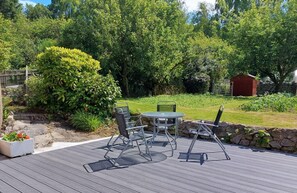 A deck with a table and chairs, surrounded by a garden with various plants and a red shed in the background. - 2 Bed in Dalbeattie (oc-d546078) (Dalbeattie)