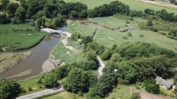 Een landelijk landschap met een brug over een rivier, omringd door begroeiing en velden.