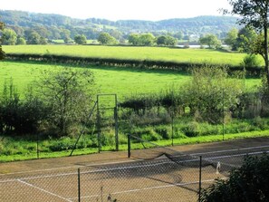 Een tennisbaan met een hek van gaas en een uitzicht op een groen veld en bomen op de achtergrond.