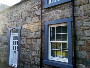 A stone building with two windows, one with a white door and the other with a sign indicating a two-bedroom self-cater accommodation. - 2 Bed in New Galloway (oc-d550751) (New Galloway)