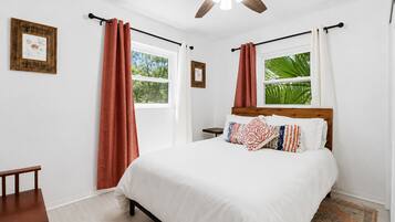 A bedroom with a bed, curtains, a ceiling fan, and a window with a view of greenery.