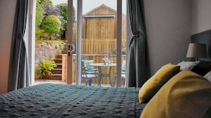 A bedroom with a bed, a window with curtains, and a view of an outdoor patio with a table and chairs. - 1 Bed in Carsethorn, Dumfries (oc-d642347) (Carsethorn)