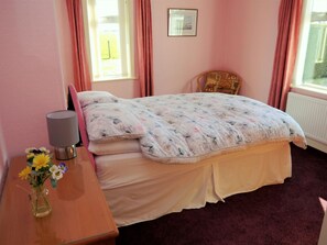 Ein Schlafzimmer mit einem Bett, einem Nachttisch mit Lampe, einem Sessel und einem Fenster mit Vorhängen.