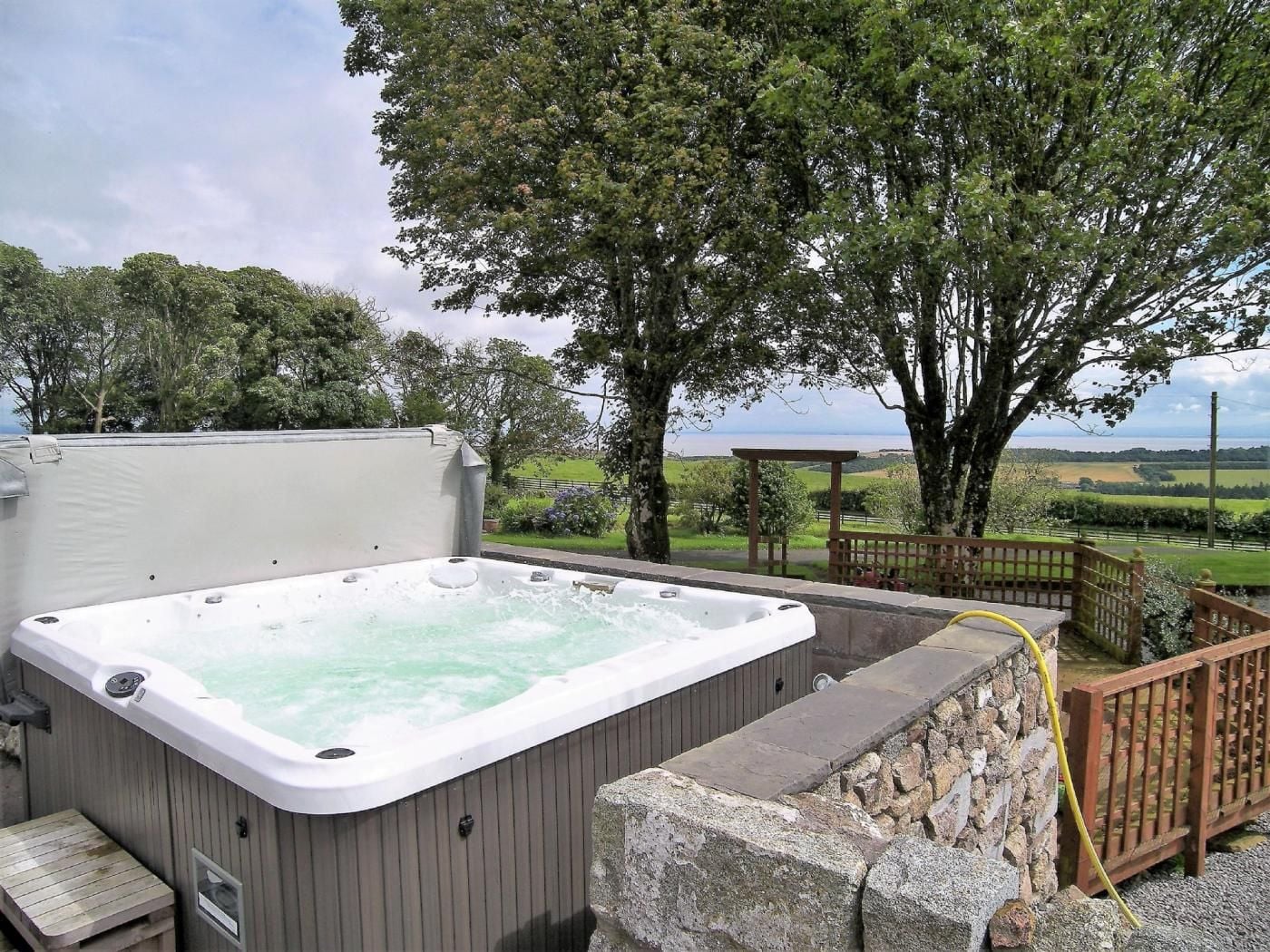 A hot tub with a stone wall and wooden deck, surrounded by trees and a view of the countryside.