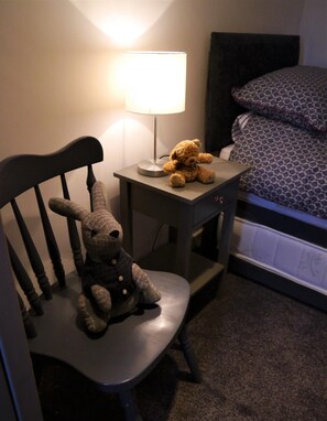 A small bedside table with a lamp, a chair with a stuffed toy, and a bed with patterned bedding. - 2 Bed in Kippford (oc-d564011) (Kippford)