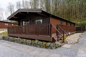 A cabin with a wooden deck, stone foundation, and a forest backdrop. - 3 Bed in Kippford (oc-d651620) (Kippford)