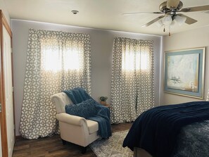 Un dormitorio con una cama, una silla, un ventilador de techo y cortinas estampadas.