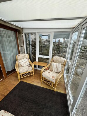 A room with two wicker chairs, a small table, and a view of a stone wall through large windows. - 2 Bed in Twynholm, Kirkcudbright (oc-d658490) (Kirkcudbright)