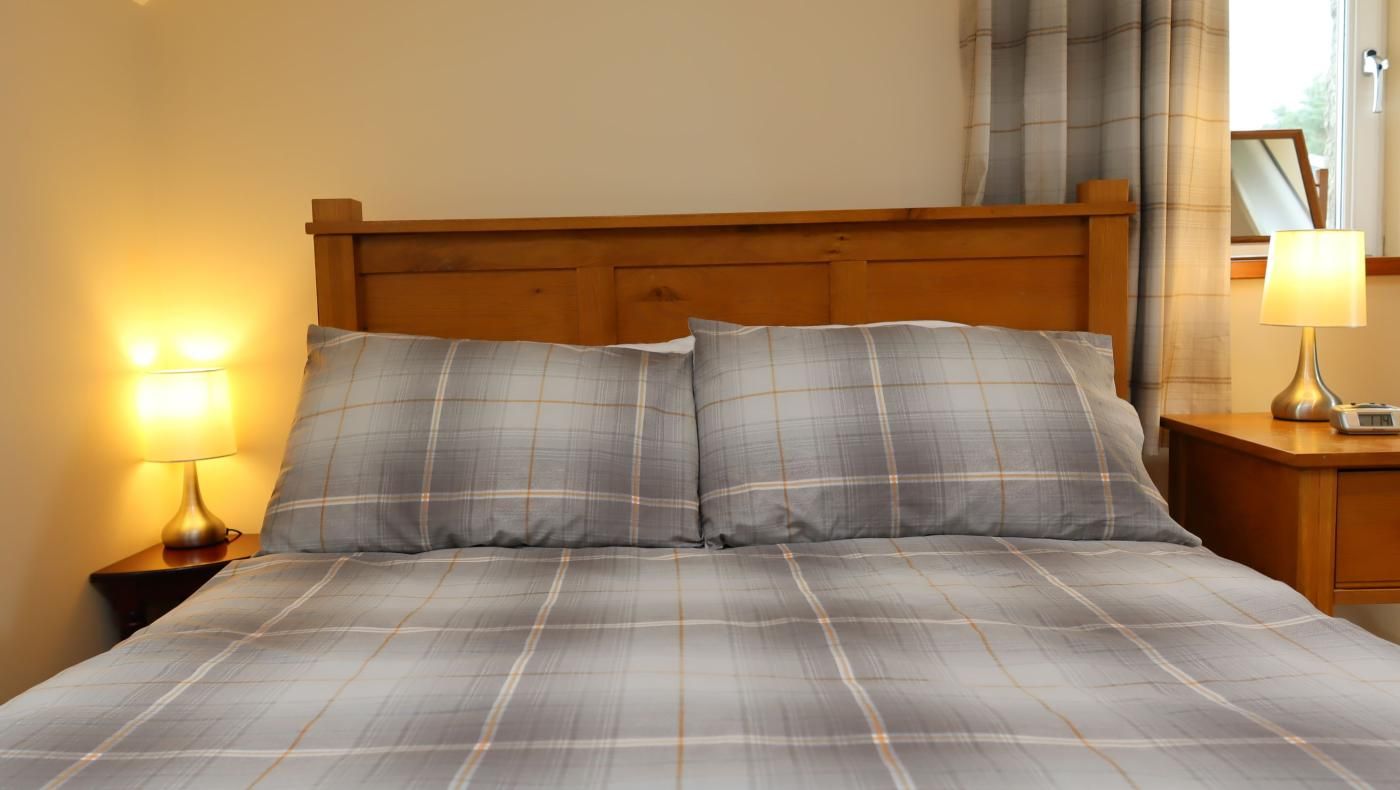 A bed with a plaid bedspread and two pillows, a nightstand with a lamp, and a window with curtains.