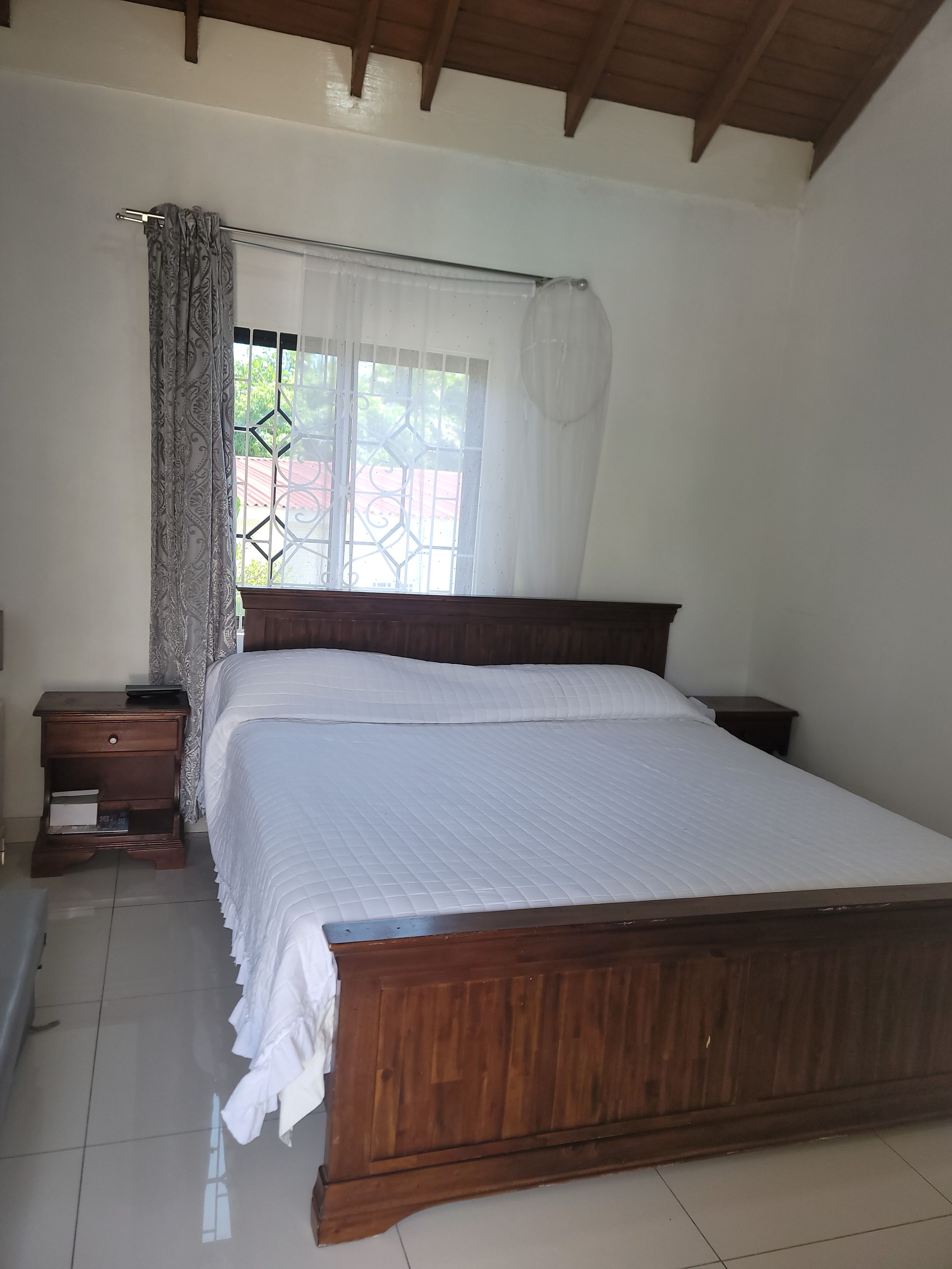 A bedroom with a wooden bed, a nightstand, a window with a curtain, and a view of greenery outside.