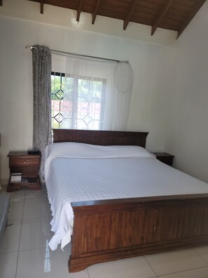 A bedroom with a wooden bed, a nightstand, a window with a curtain, and a view of greenery outside. - Cozy 2-bedroom apartment with WIFI AND AC in charming Tower Isle (Tower Isle)