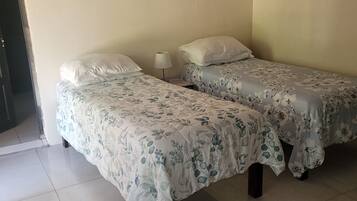 A room with two beds, each with floral bedding, a bedside table with a lamp, and a mosquito net hanging on the wall.