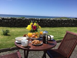 Un coin petit-déjeuner aménagé avec un mur en pierre et une vue sur l’océan, comprenant une table avec des croissants, des fruits, du café et un vase de fleurs.
