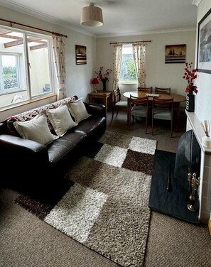 A living room with a black leather sofa, a dining table with chairs, a fireplace, and a large window with curtains. - 3 Bed in Newton Stewart (oc-d545503) (Port William)