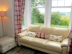 A beige leather sofa with matching cushions, a small white chest of drawers, and a window with a view of greenery. - 4 Bed in Castle Douglas (oc-d615180) (Corsock)