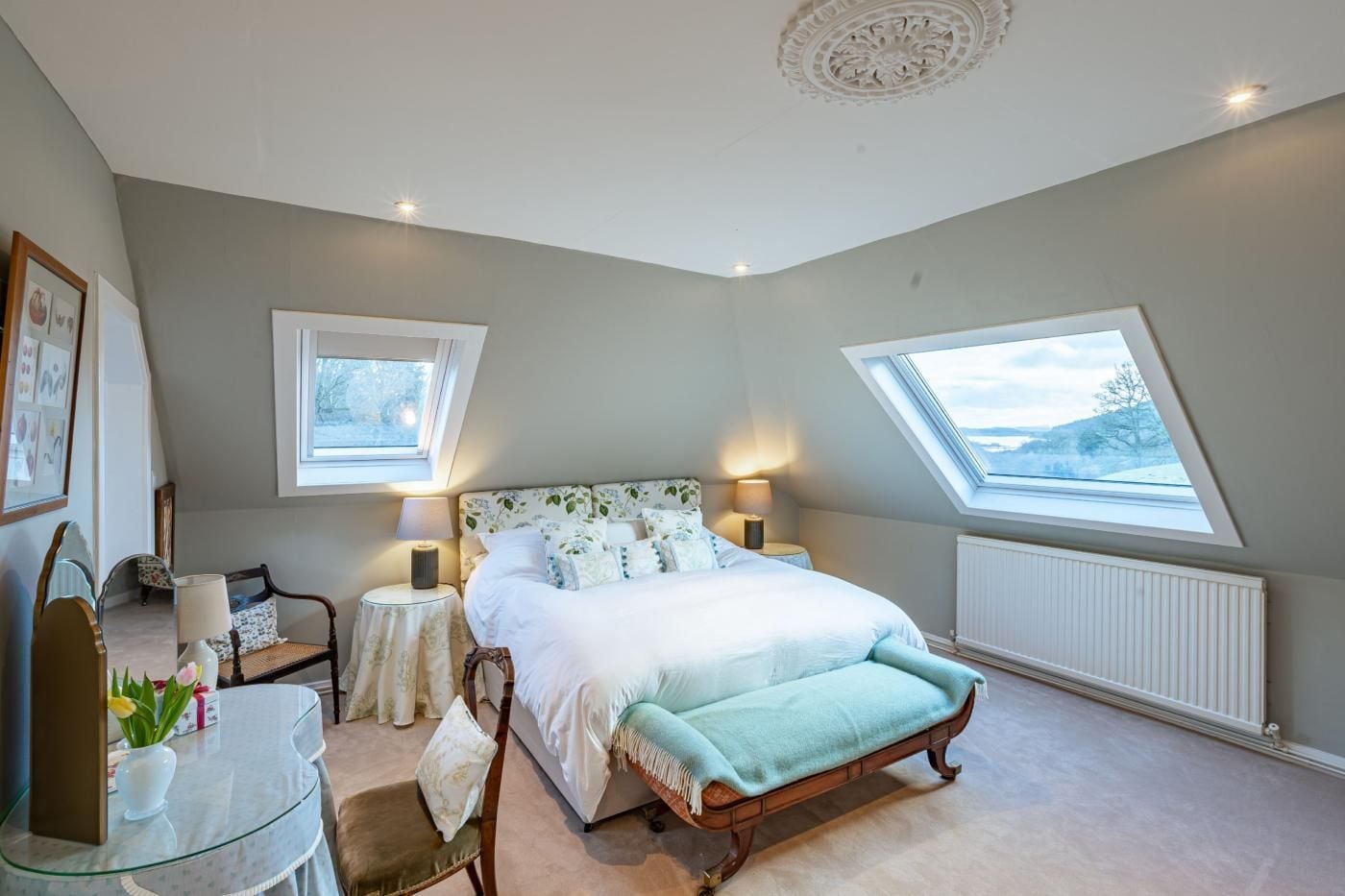 A bedroom with a bed, two bedside tables, a chair, a small table, and two windows.