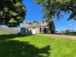Une maison en pierre avec une grande pelouse devant, une zone clôturée sur la droite et une colline en arrière-plan.