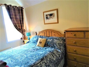 Un dormitorio con cama, cómoda, mesita de noche, lámpara, ventana con cortinas y un cuadro en la pared.