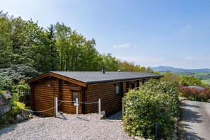 A wooden cabin with a gravel driveway, surrounded by greenery and a clear sky. - 3 Bed in Kippford (oc-d597989) (Kippford)