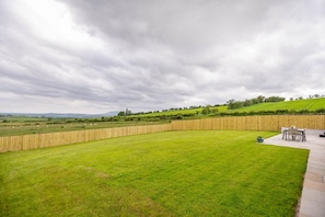 A well-maintained grassy area with a wooden fence, a patio with outdoor furniture, and a view of rolling hills in the distance. - 3 Bed in Dumfries (oc-d641667) (Torthorwald)