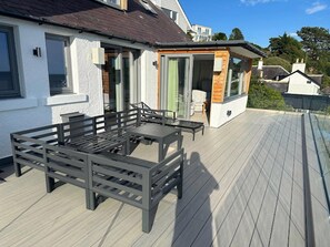 Eine Terrasse mit Tisch und Stühlen, eine Glastür, die zu einem Raum führt, und ein weißes Gebäude mit dunklem Dach.
