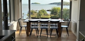 Ein Essbereich mit Tisch und Stühlen, große Fenster mit Blick auf das Meer und viel Grün.
