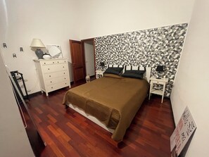 A bedroom with a bed, a dresser, a nightstand, a lamp, and a geometric-patterned wall. - Experience Genoa: A Charming Apartment in the Heart of Its Historic Center (Genova)