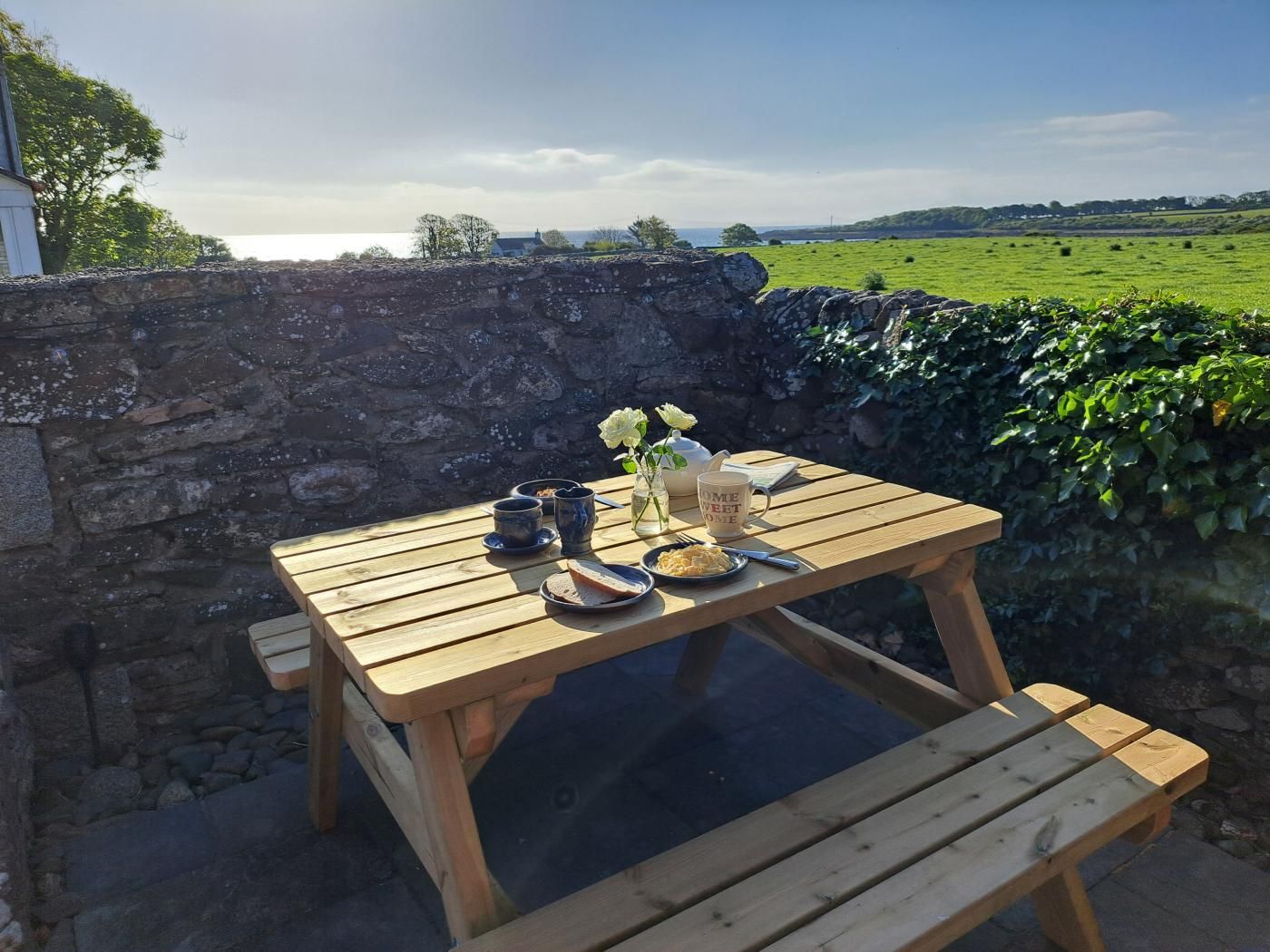 Ein Holztisch mit einer Glasplatte draußen, umgeben von einer Steinmauer, mit Blick auf ein grünes Feld und ein Gewässer in der Ferne.
