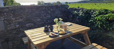 Ein Holztisch mit einer Glasplatte draußen, umgeben von einer Steinmauer, mit Blick auf ein grünes Feld und ein Gewässer in der Ferne.