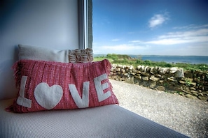Un coussin orné du mot « AMOUR », posé sur un rebord de fenêtre, d’où l’on aperçoit un mur de pierre et de la verdure.