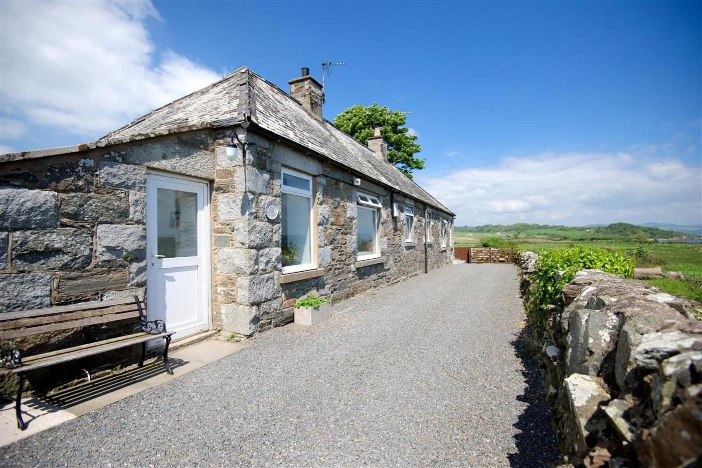A stone cottage with a gravel driveway, a bench, and a stone wall.