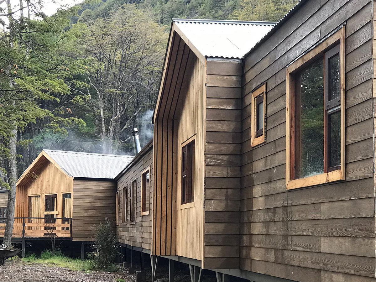 A wooden cabin with a metal roof, a chimney emitting smoke, and a small deck area.