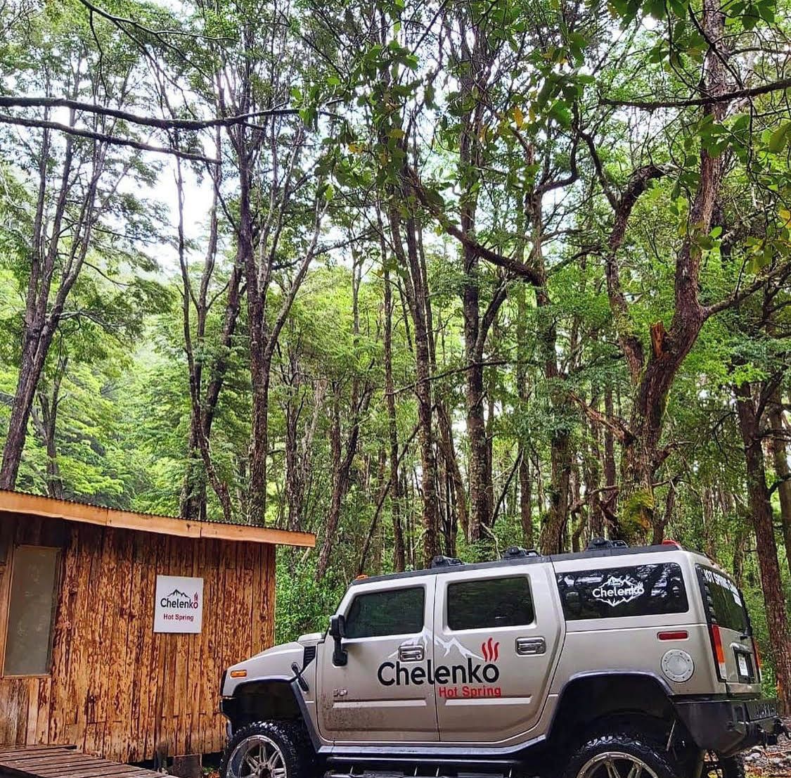 A vehicle parked in a forested area with a wooden structure labeled 'Chelenko Hot Spring' in the background.