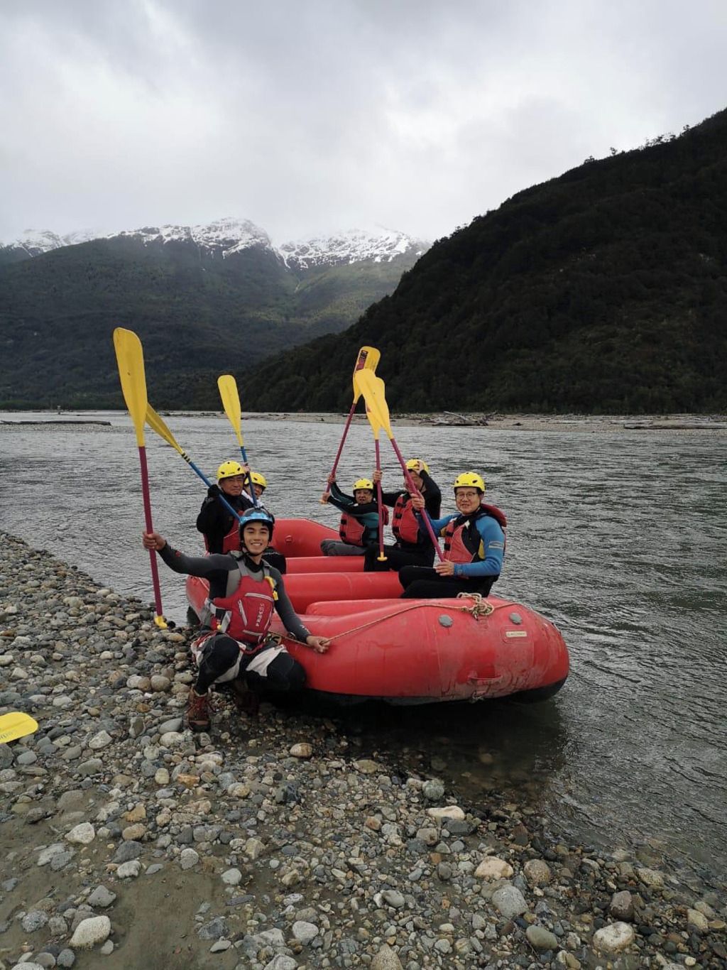 Un groupe de personnes se trouve dans un radeau gonflable rouge, sur une plage rocheuse au bord d’un lac, avec des montagnes en arrière-plan.