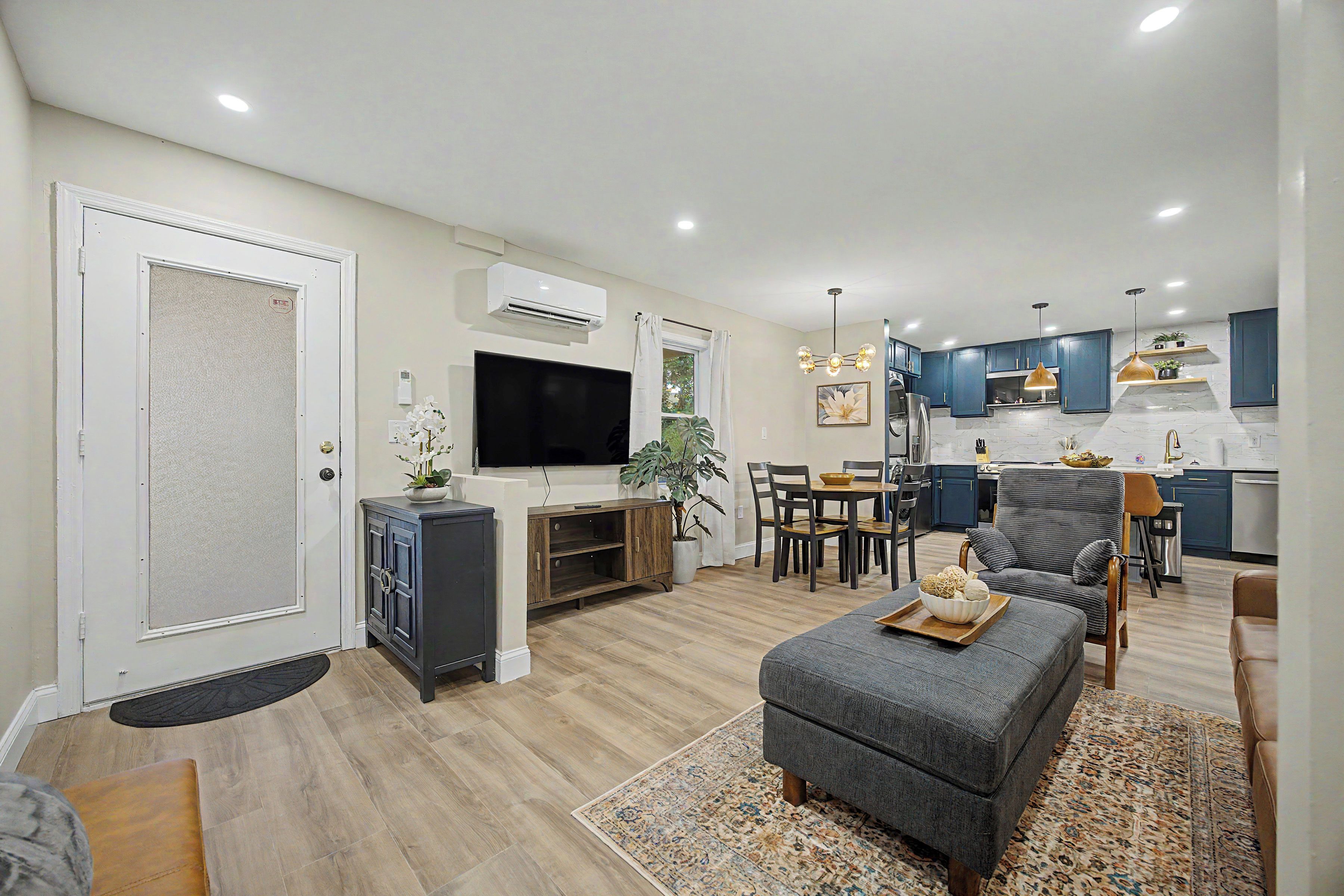 A modern living room with a flat-screen TV, a dining area with a wooden table and chairs, and a kitchen with blue cabinets and a stainless steel refrigerator.