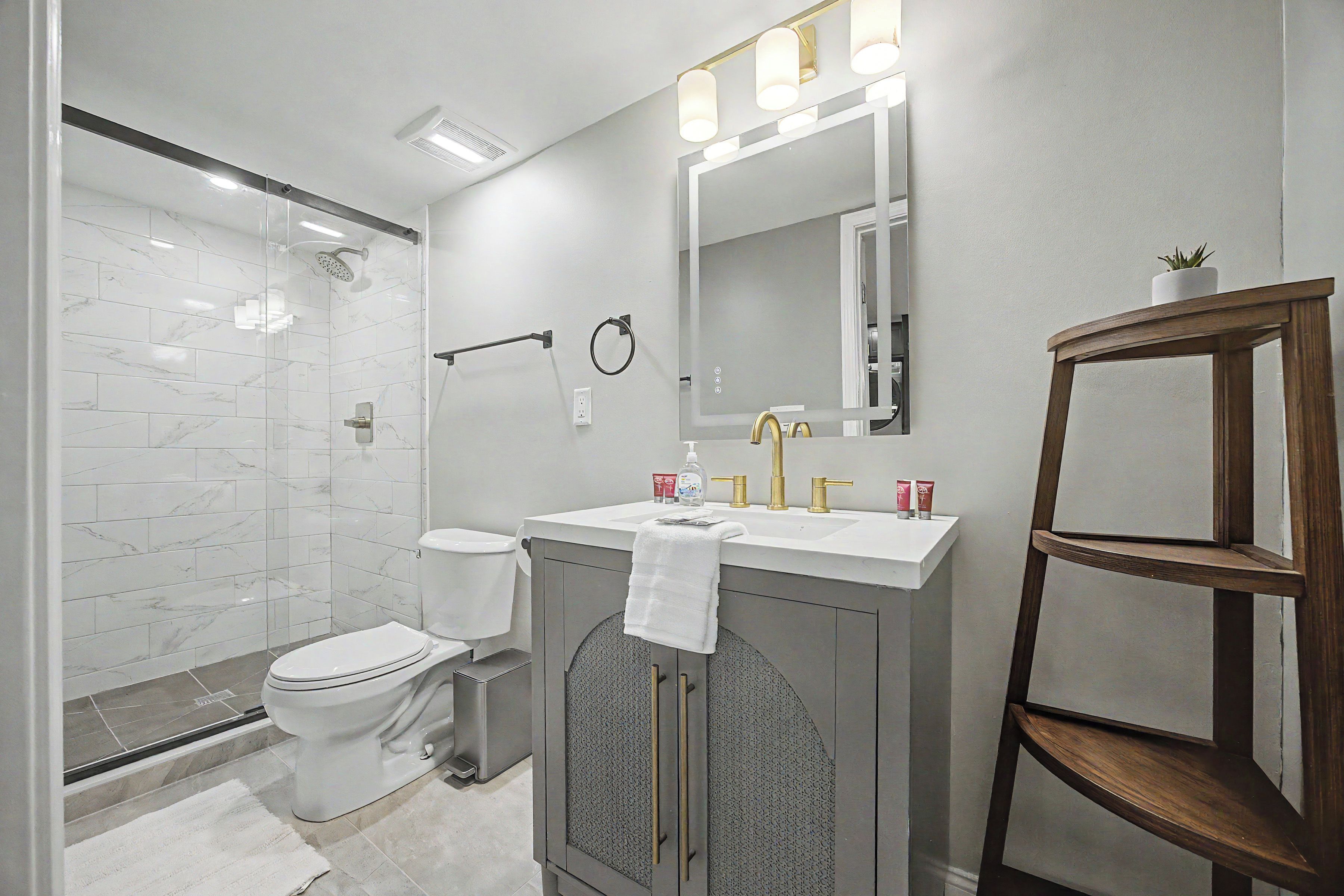 A modern bathroom with a glass-enclosed shower, a toilet, a vanity with a mirror, and a wooden shelf.