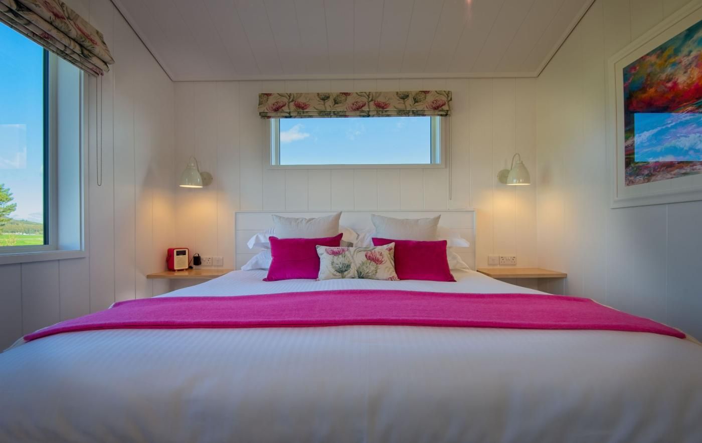 Uma cama com colcha branca e rosa, duas luminárias de cabeceira, uma mesinha de noite com um rádio e uma janela com vista para o verde.
