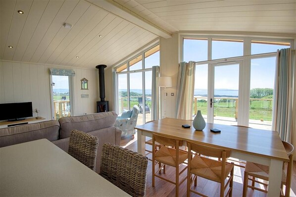 A spacious living area with a sofa, a television, a table with chairs, and large windows offering a view of the outdoors. - 3 Bed in Mossyard, Gatehouse Of Fleet (oc-d546163) (Gatehouse of Fleet)