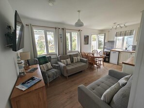 Ein Wohnzimmer mit einer Couch, einem Esstisch mit Stühlen, einer Küchenzeile und großen Fenstern mit Vorhängen.