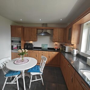 A kitchen with wooden cabinets, a white dining table with blue chairs, and a window providing natural light. - 4 Bed in Southgate (oc-n46057) (Southgate)