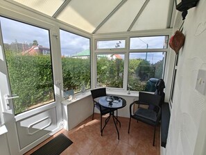 Une véranda avec une table et des chaises, de grandes fenêtres et une vue sur la verdure et les bâtiments.