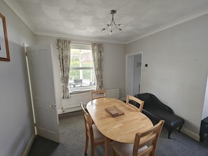 Une salle à manger avec une table ronde en bois, quatre chaises, un canapé noir et une fenêtre avec des rideaux.