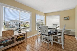 Un coin repas avec une table et des chaises, de grandes fenêtres équipées de stores, un petit meuble contenant des livres et un radiateur.