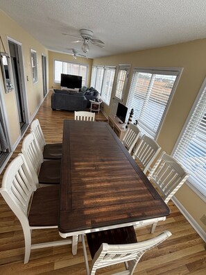 A long dining table with white chairs, a ceiling fan, and large windows with blinds. - Beautiful Cherry Grove Beach House (North Myrtle Beach)