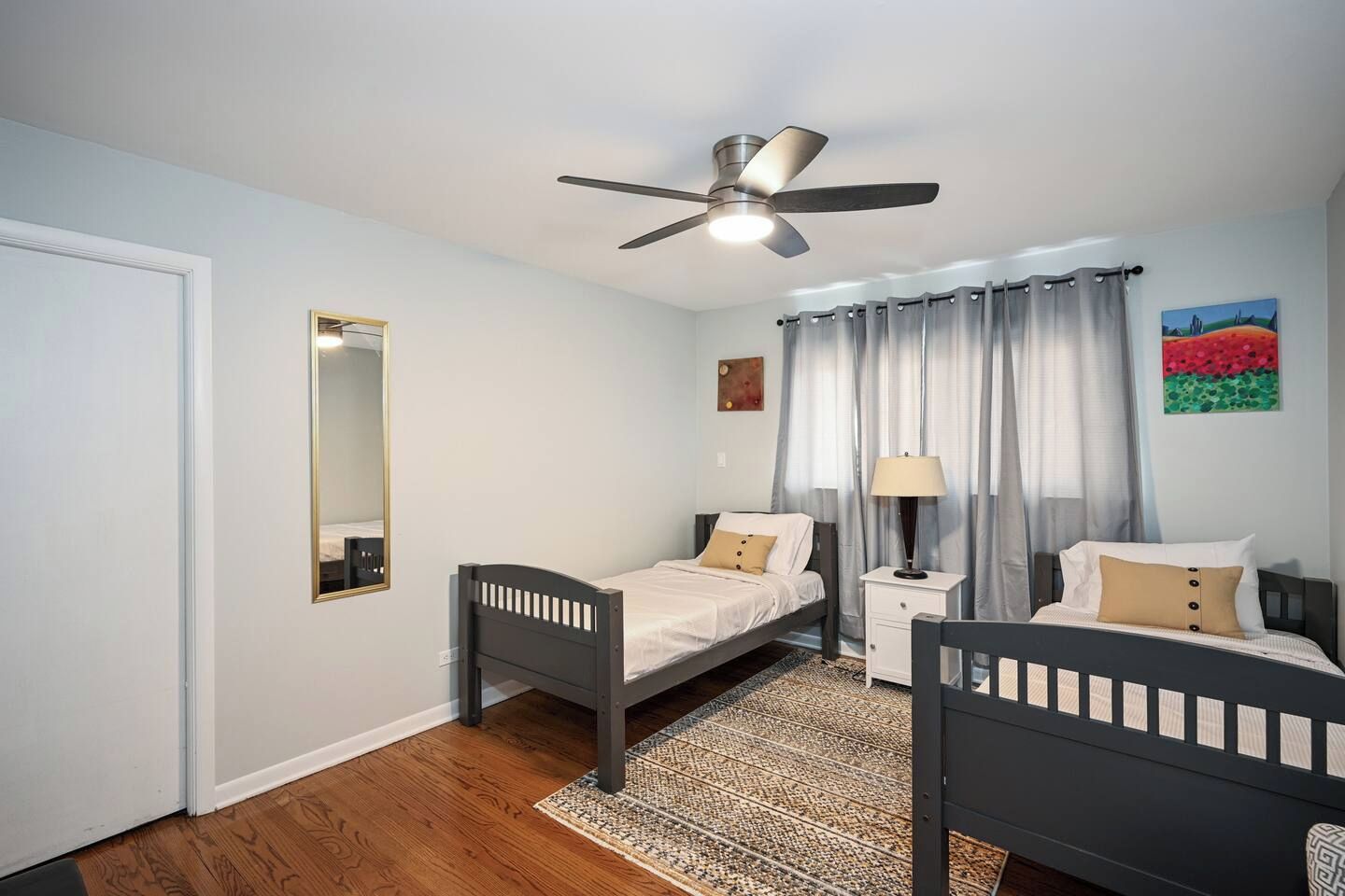 A bedroom with two single beds, a ceiling fan, a bedside table with a lamp, a mirror, and a painting on the wall.
