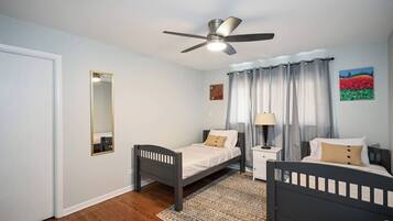 A bedroom with two single beds, a ceiling fan, a bedside table with a lamp, a mirror, and a painting on the wall.