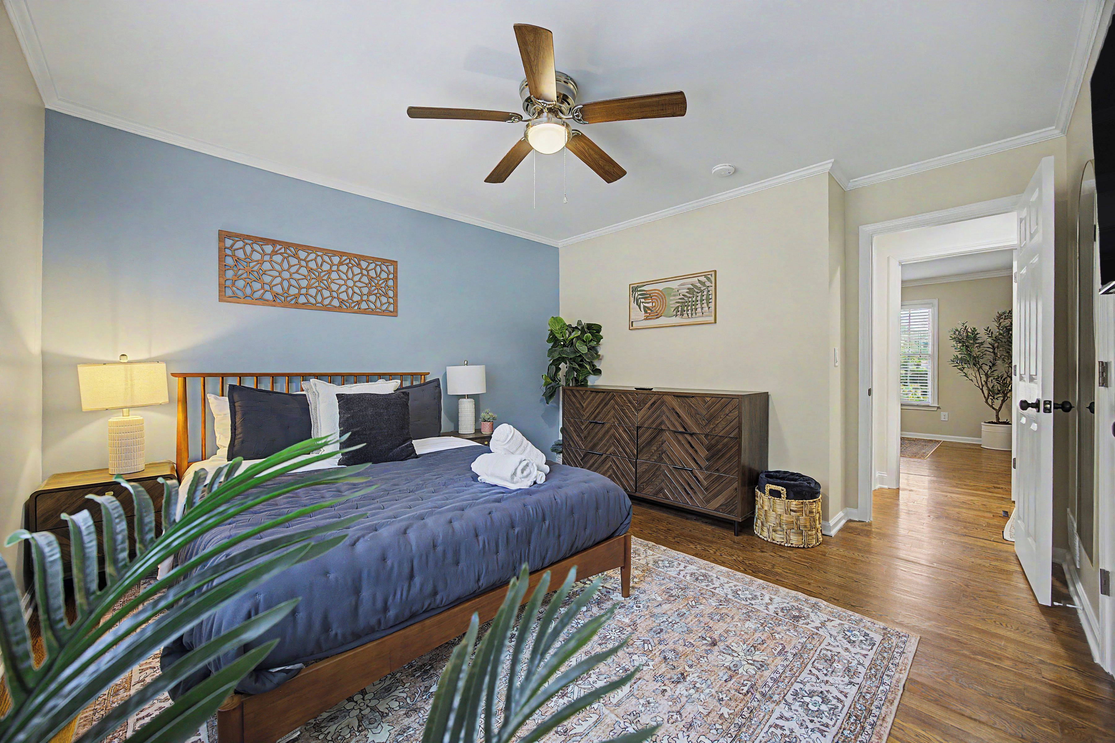 A bedroom with a bed, a ceiling fan, a nightstand, a lamp, a decorative wall piece, a potted plant, and a door leading to another room.